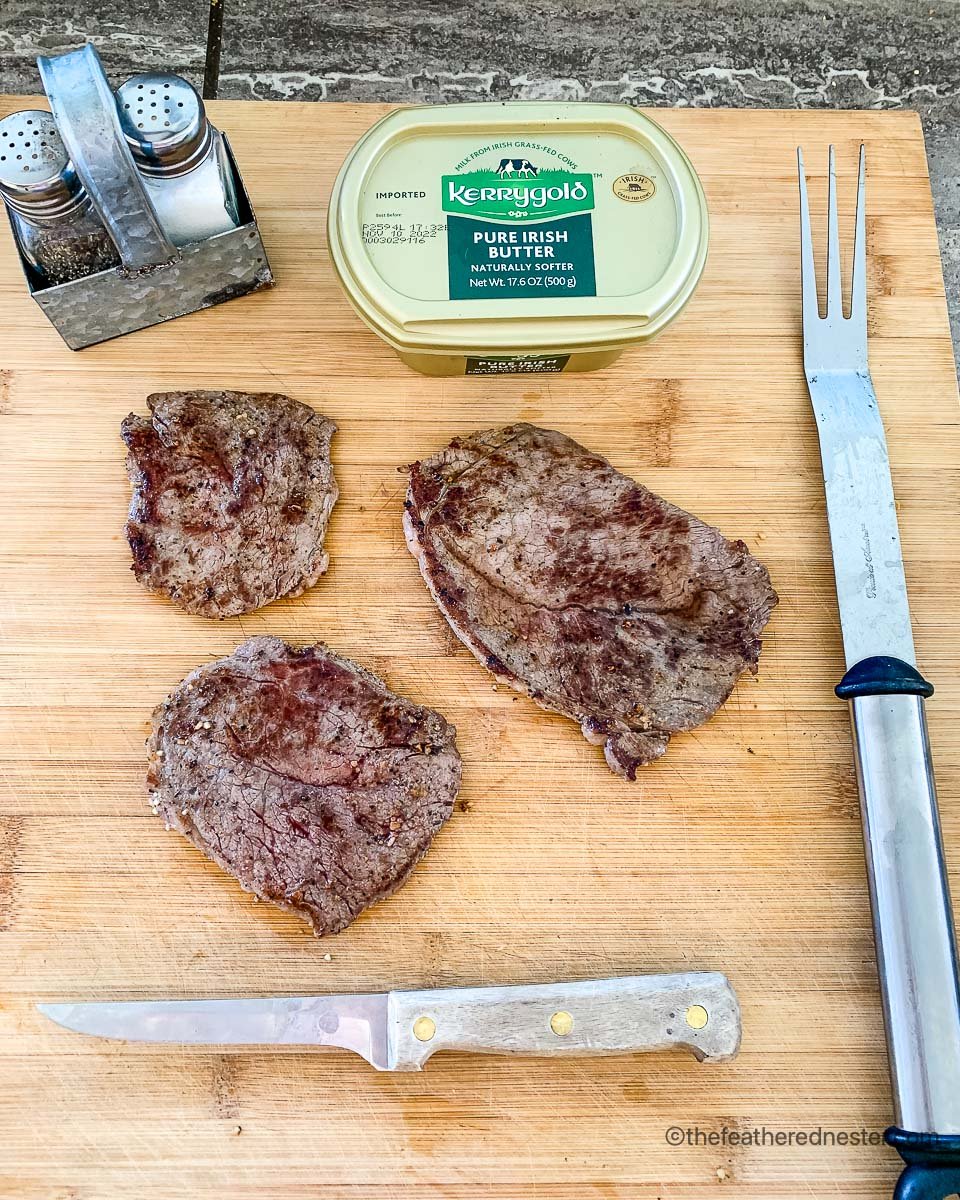 Cooked Blackstone Griddle Steaks on a wood board with Butter, Salt, Pepper, Knife and a Fork on the side. 