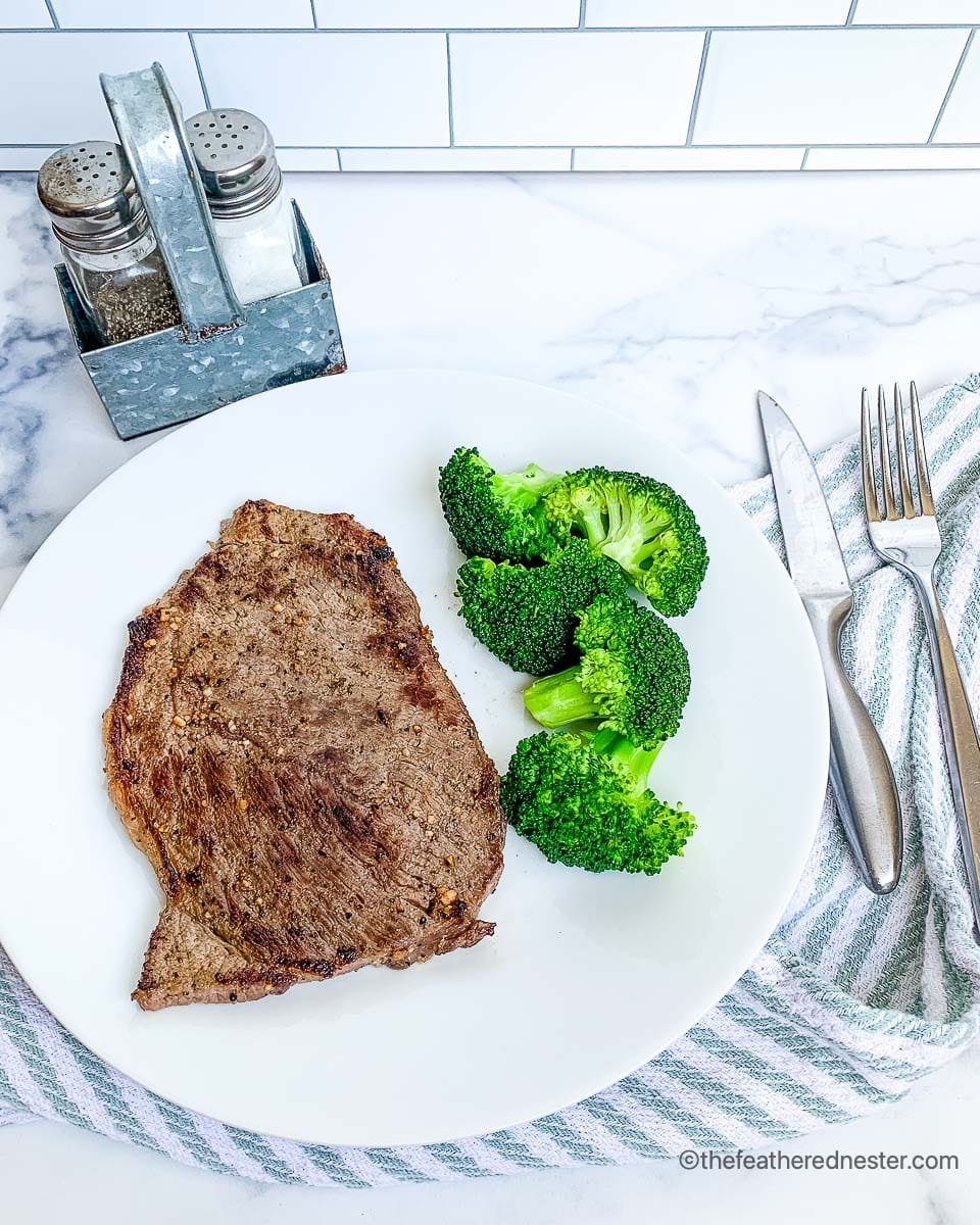 cooked steak on a blackstone griddle with broccoli on a white plate with silverware and a blue and white napkin.