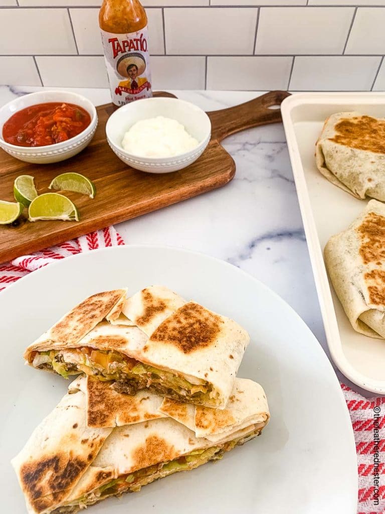 homemade crunch wrap supremes on a white plate with a serving board with bowls of sour cream, salsa, a bottle of Tapatio, and lime slices in the background