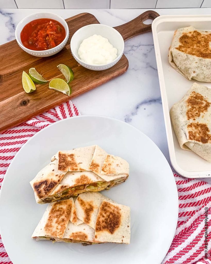 crunchwrap cut on half on a white plate with a tray of another one on a tray and a serving board with bowls of sour cream, salsa, and lime slices in the background
