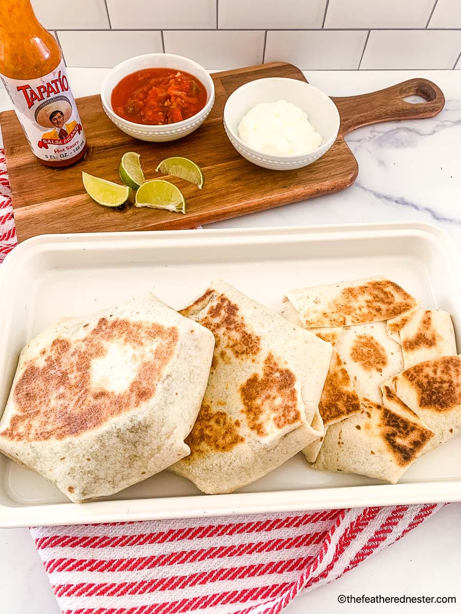 blackstone crunchwrap on a white plate with a serving board with bowls of sour cream, salsa, a bottle of Tapatio, and lime slices in the background.