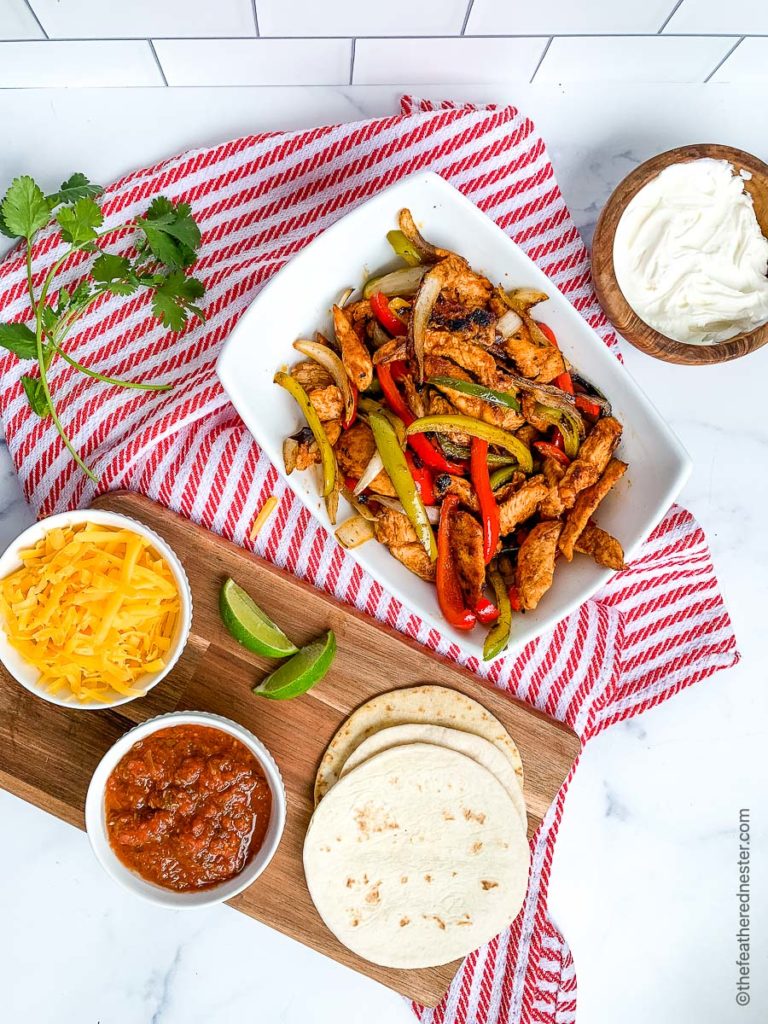 a white bowl of chicken fajitas with bowls of shredded cheese, salsa, sour cream, and a stack of flour tortillas