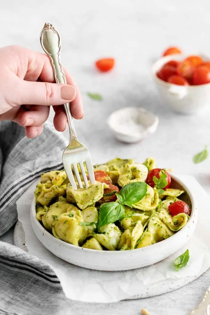 a hand using silverware in a white bowl of tortellini caprese salad with tomatoes and salt in the background.