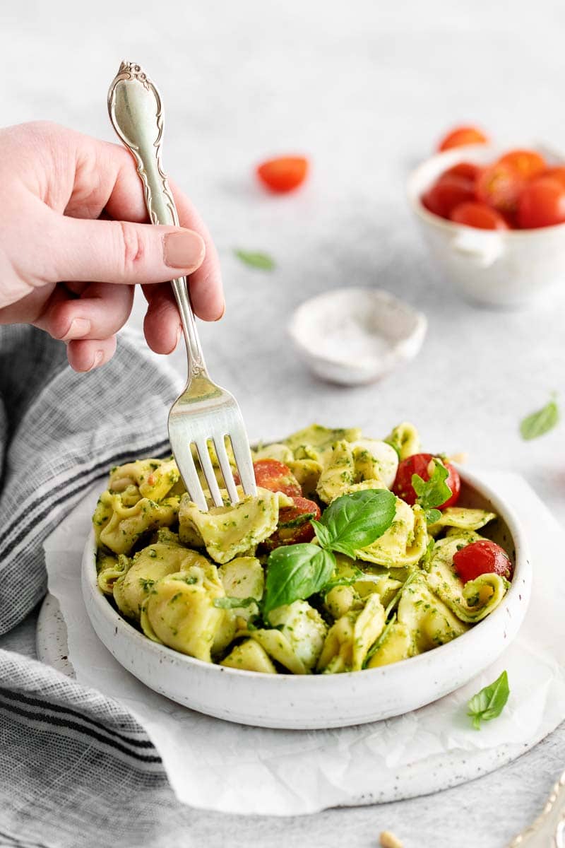 Hand holding a fork in a bowl of tortellini wtih pesto sauce.