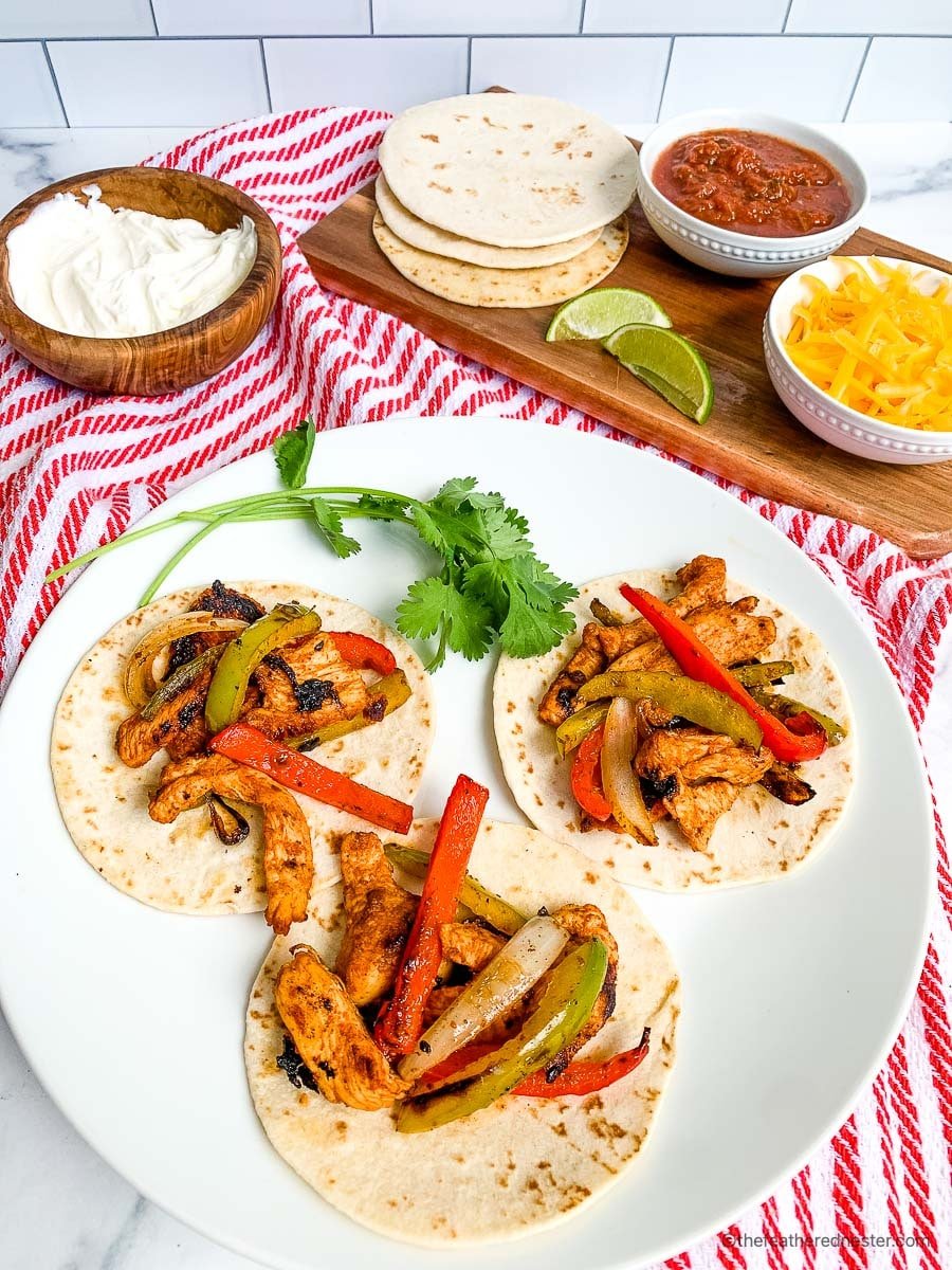 a plate of three chicken fajitas on flour tortilla on a white plate with a sprig of cilantro on top of a red and white napkin.