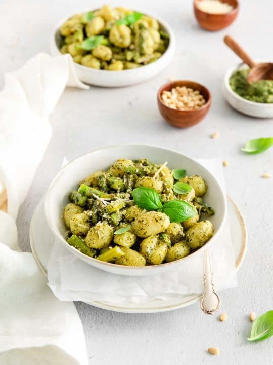 two white bowls of pesto gnocchi asparagus with small bowls of grated parmesan cheese in the background