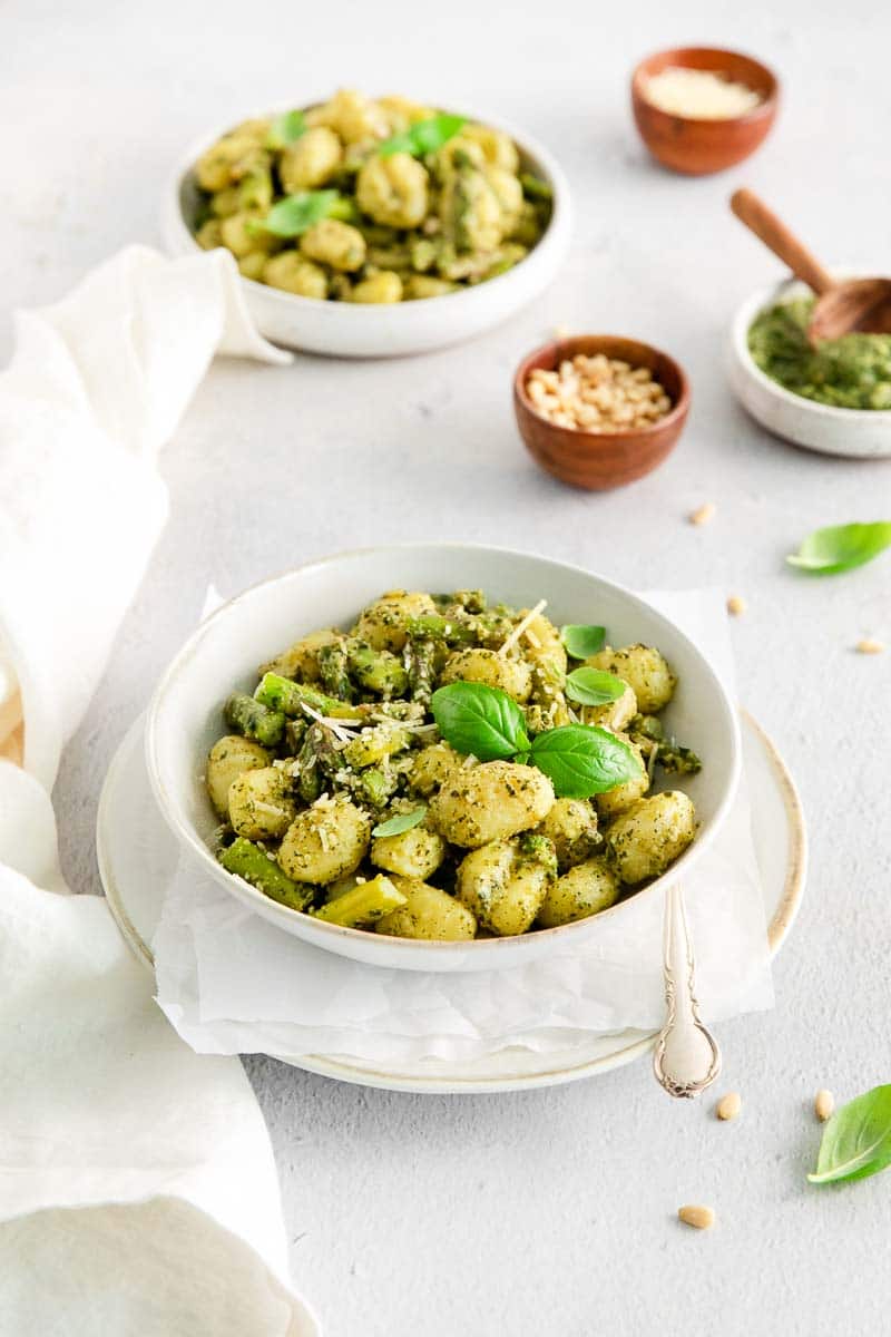 two white bowls of pesto gnocchi asparagus with small bowls of grated parmesan cheese in the background.