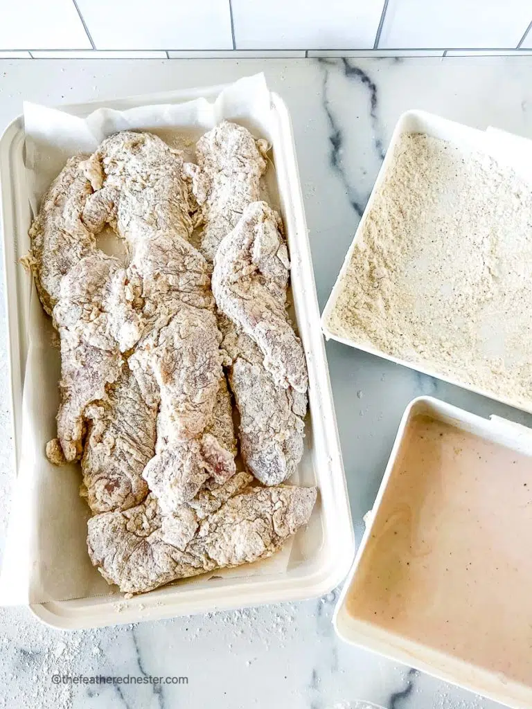 Chicken tenders that are coated in flour and buttermilk mixture on a white serving platter along with flour mixture and buttermilk mixture on separate trays