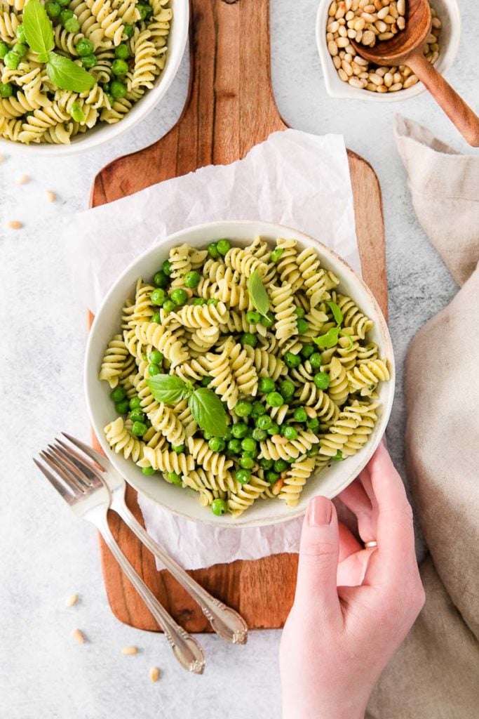 a hand holding a plate of rotini pasta salad with green vegetables and silverware beside the plate.