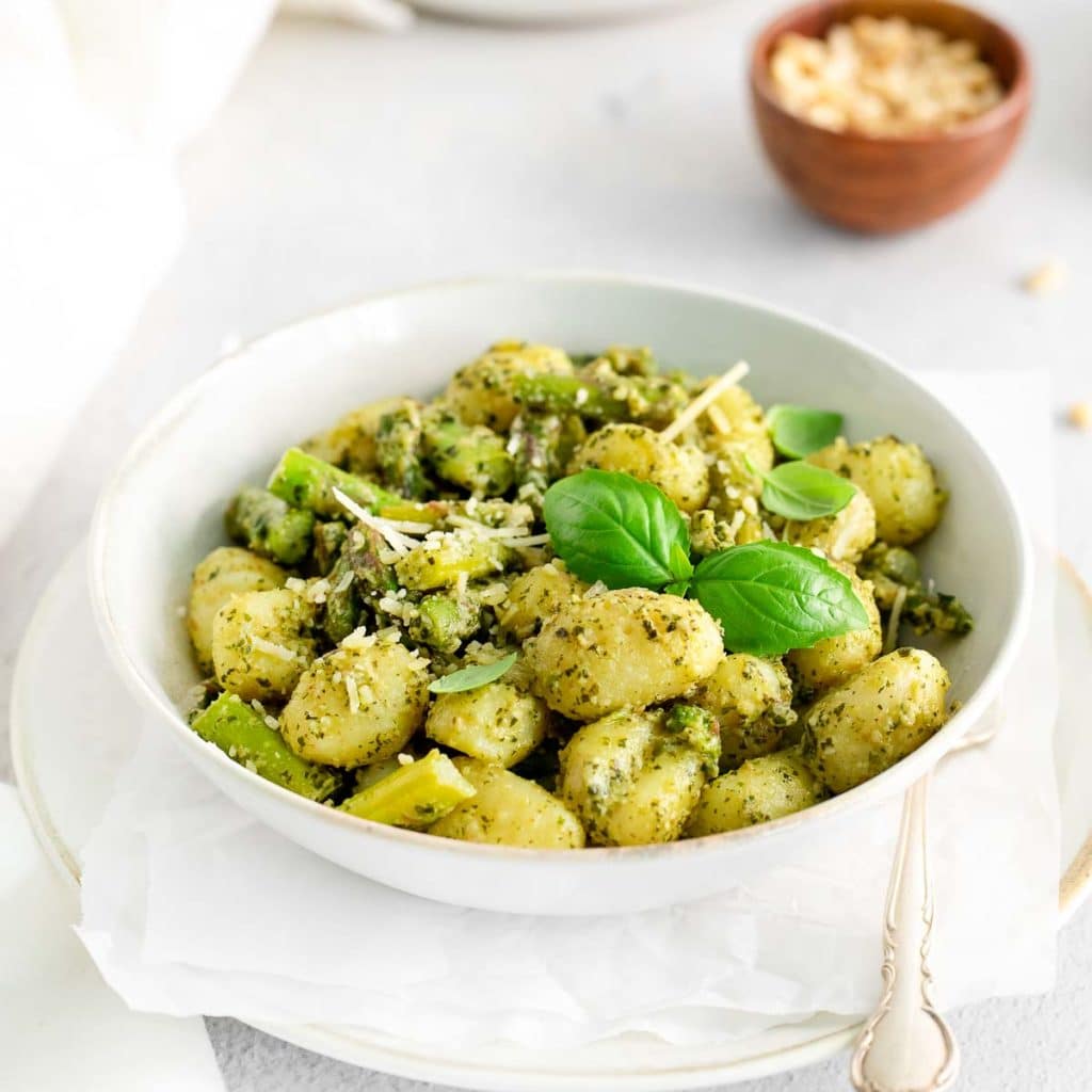 a white bowl of pesto gnocchi with asparagus with a small bowl of parmesan cheese in the background