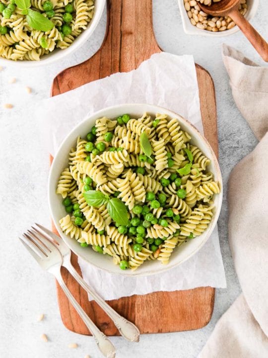 light summer meal of pasta with peas on a wood serving board with silverware and dish of pine nuts.