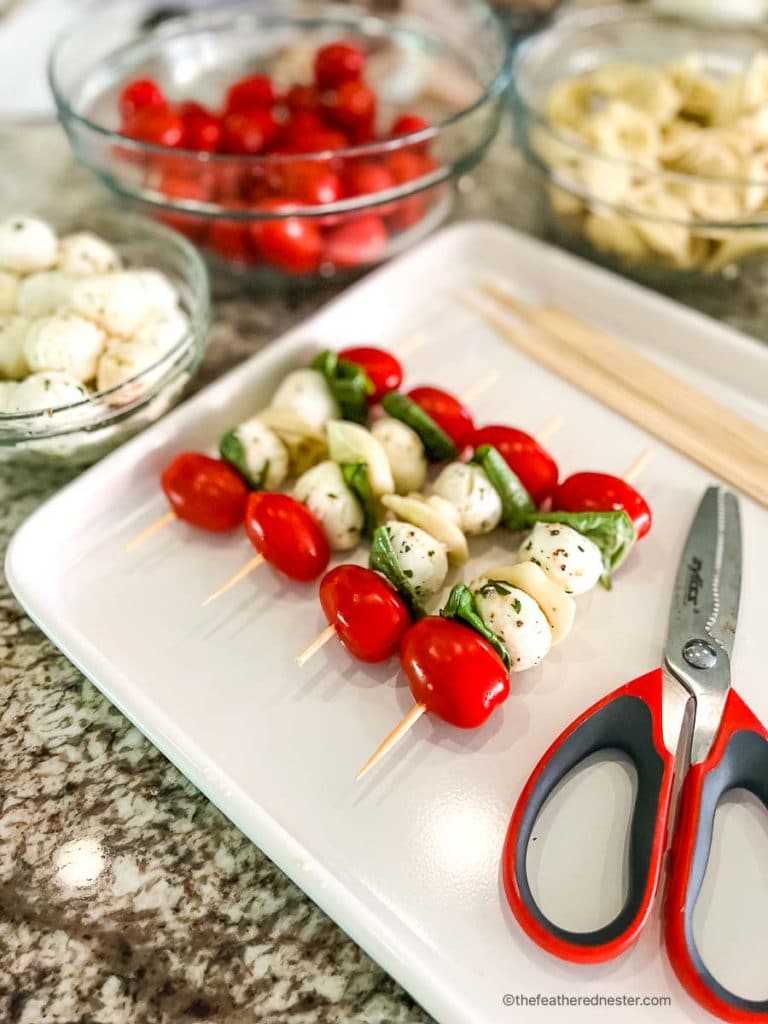 bowls of cherry tomatoes, mozzarella balls, pasta, and basil leaves along with a tray of 4 assembled tortellini appetizers and a pair of scissors on a white serving tray