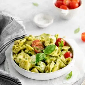 a white bowl of tortellini caprese salad with tomatoes and salt in the background