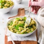 Woman's hand holding a fork in bowl of rotini pesto pasta with veggies, sitting on top of a wooden serving board.