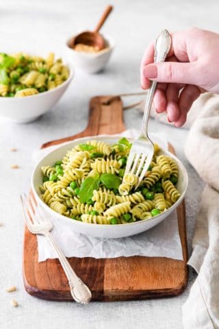 pea pesto pasta on a white salad plate on top of a wooden board with a hand holding a silver fork