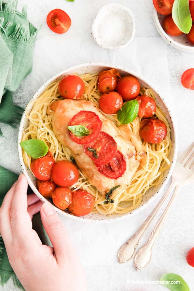 a hand holding a white bowl of pasta with a chicken breast and roasted cherry tomatoes and basil on top , ready to serve caprese chicken bowl
