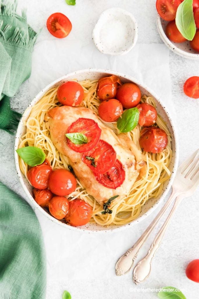 a white bowl of pasta with a chicken breast and roasted cherry tomatoes and basil on top with a white bowl of cherry tomatoes in the background