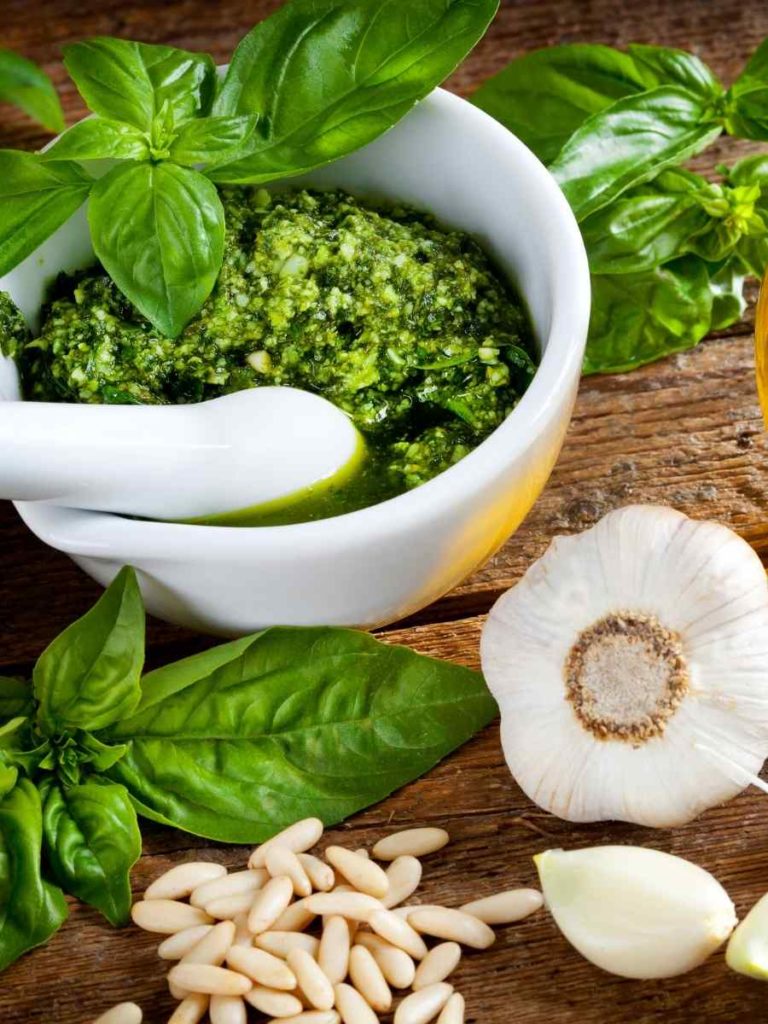 pesto sauce on a mortar and pestle with basil leaves, pine nuts, and garlic in the background