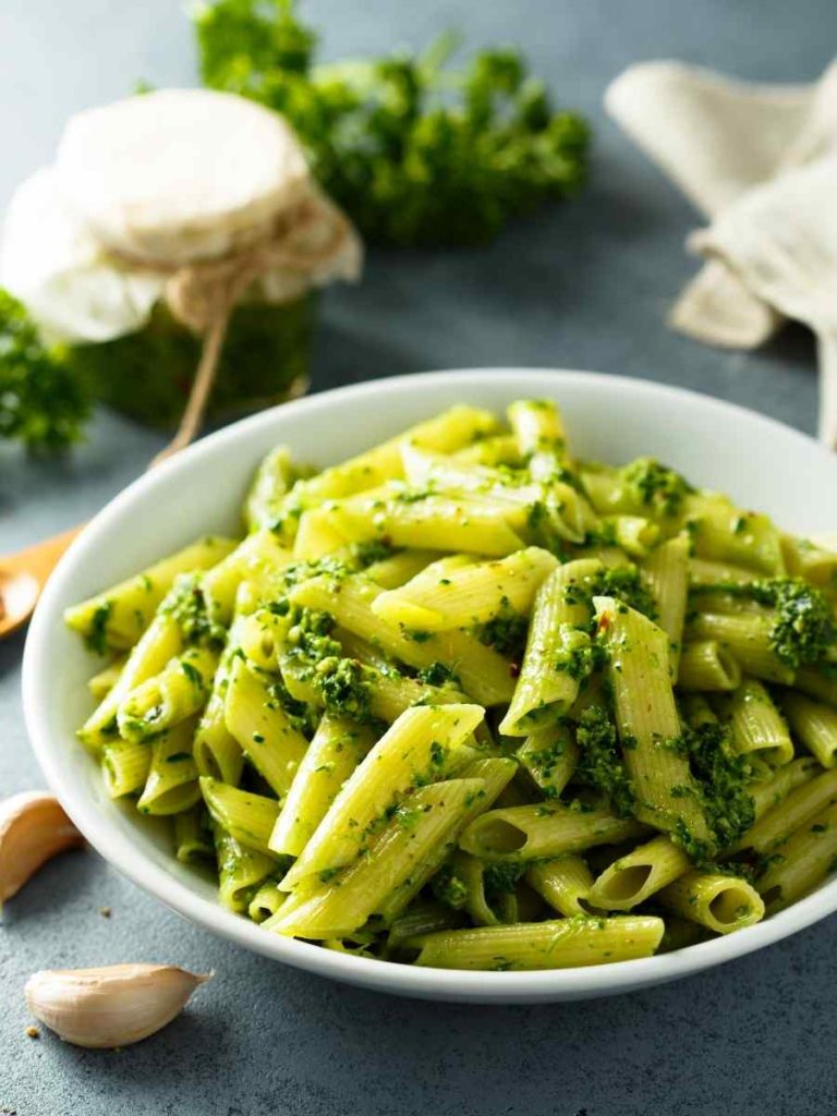 pesto penne on a white bowl with herbs, garlic, and pesto sauce on a jar in the background