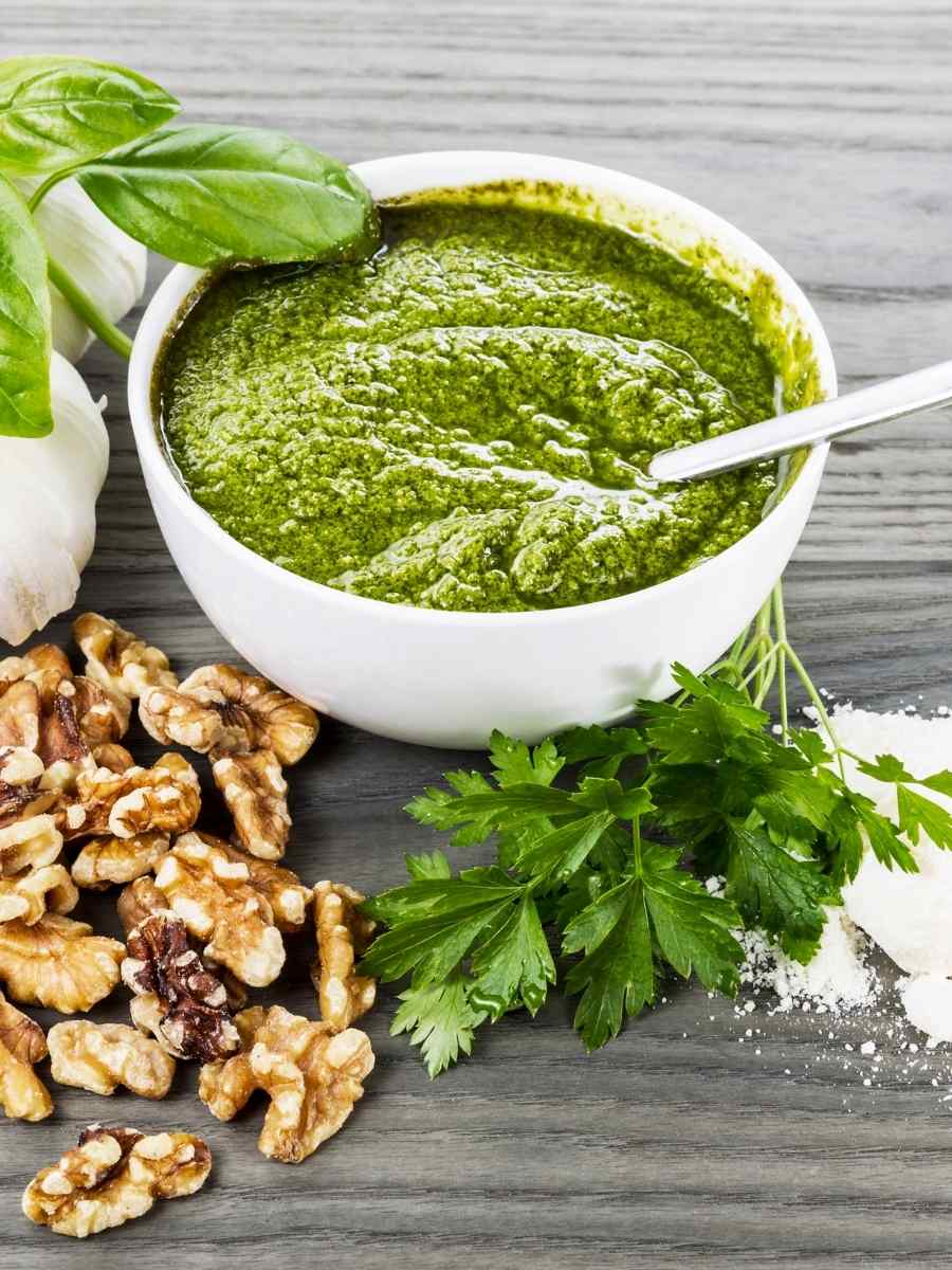 pesto sauce in a white bowl with walnuts, garlic, basil and parsley in the background