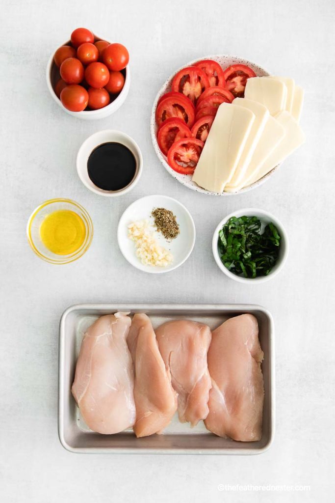 the ingredients for caprese chicken: a baking sheet with chicken breasts, white bowls of Roma tomatoes, cherry tomatoes, mozzarella cheese, olive oil, balsamic glaze, seasonings, and basil leaves