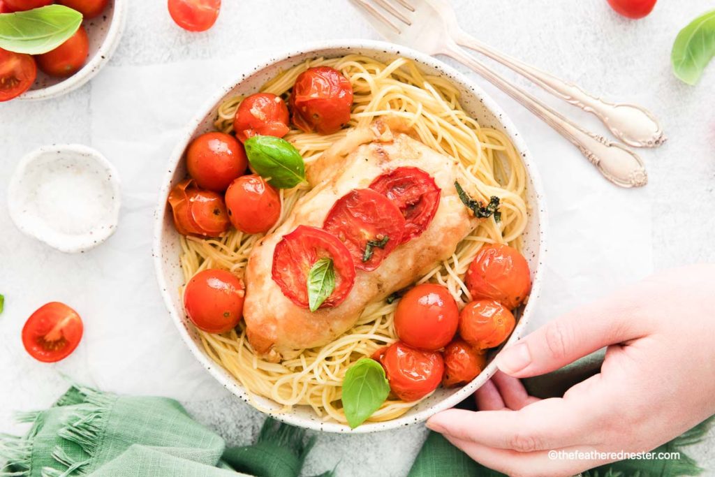 a hand holding a white bowl of pasta and chicken with roasted tomatoes