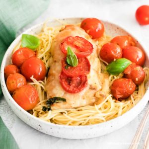a white bowl of pasta with a chicken breast and roasted cherry tomatoes and basil on top with a white bowl of cherry tomatoes in the background.