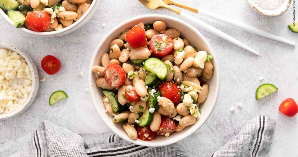 a white bowl of tuscan bean salad with silverware to the right and a striped napkin on the left with the corner of another bowl of white bean salad showing on the top left