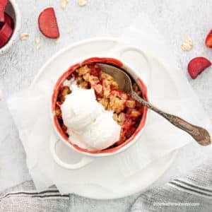 a white ramekin of apple strawberry cobbler with vanilla ice cream on top, and a spoon ready to serve it