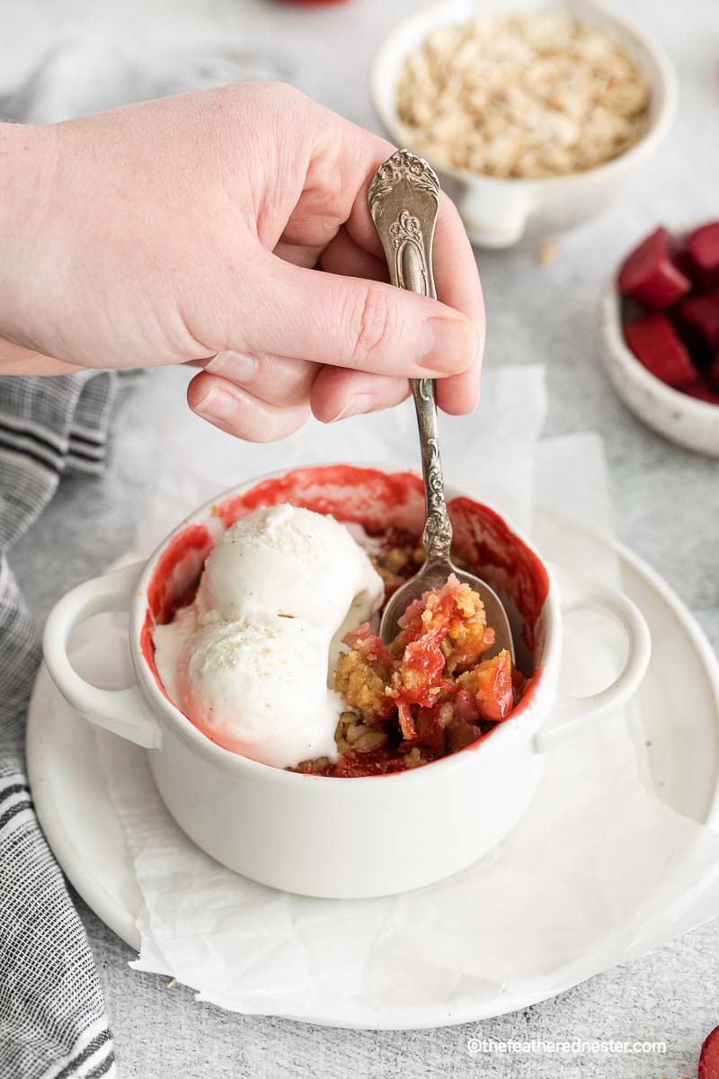 a hand holding a spoon scooping the apple strawberry crisp with vanilla ice cream on top placed in a ramekin on top of a plate with parchment paper with gray cloth, bowl of oats and rhubarb in the background.