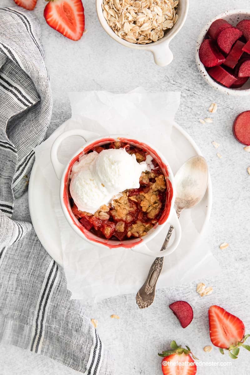 top view of a ramekin with apple strawberry crisp with vanilla ice cream on top and a spoon beside it on top of a plate with parchment paper with gray cloth, sliced strawberries, bowl of oats and rhubarb in the background.