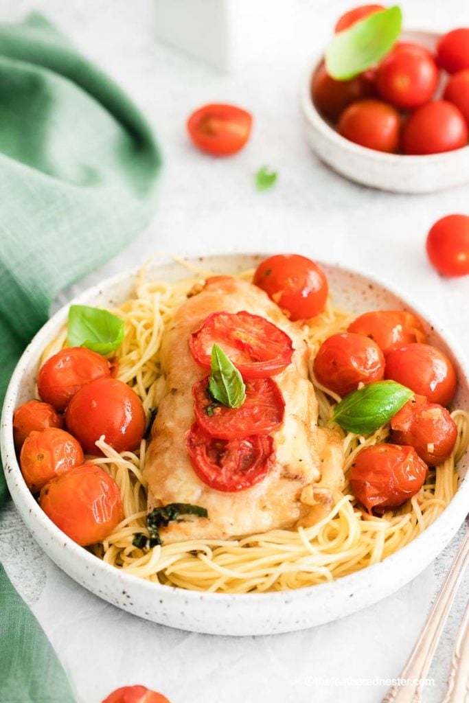 a white bowl of pasta with a chicken breast and roasted cherry tomatoes and basil on top with a white bowl of cherry tomatoes in the background