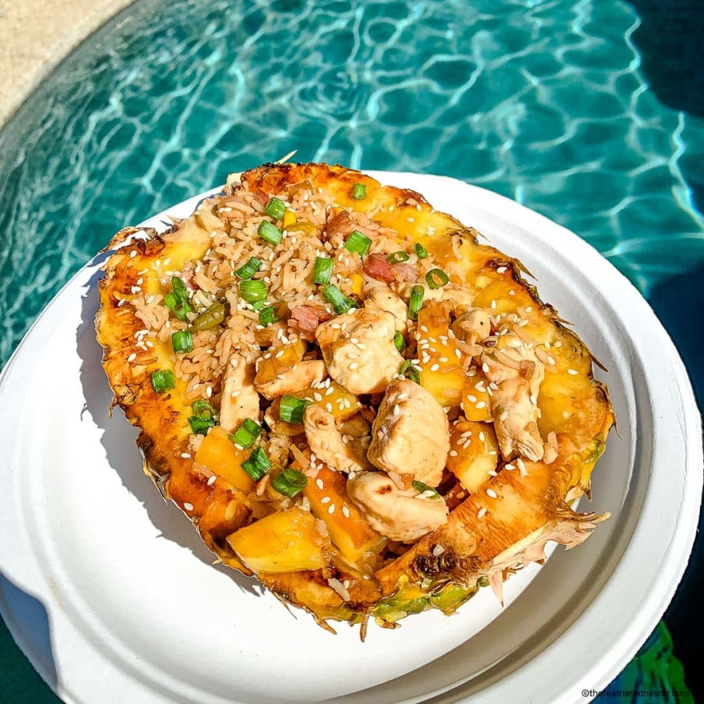 chicken teriyaki pineapple bowl on a white plate held outdoors with swimming pool water at the background