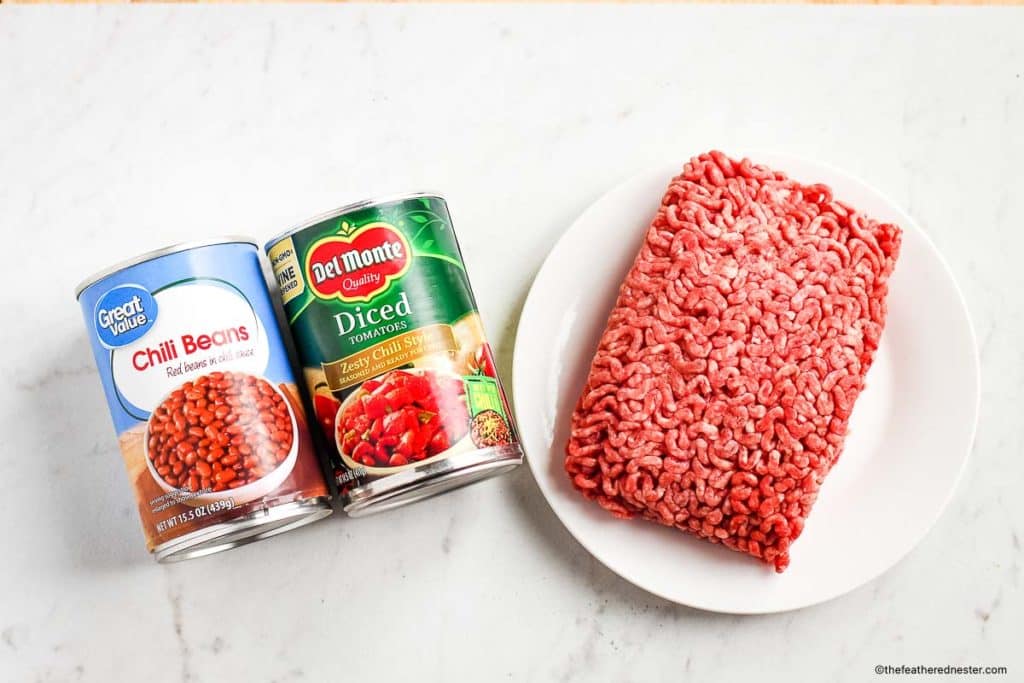 Ingredients for the stovetop chili recipe which consist of canned chili beans, canned diced tomatoes, and a ground beef on a white plate.
