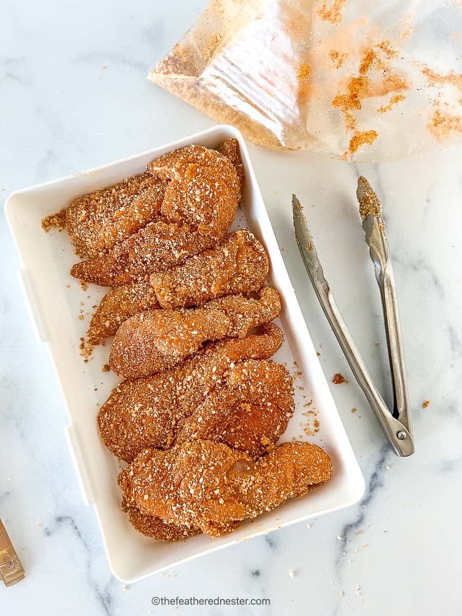 Chicken tenders coated with seasonings on a casserole with tongs at the side.