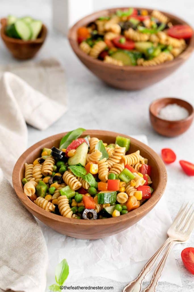 zesty Italian pasta salad and silverwares on a wood bowl with another bowl of salad and salt, cucumber, and cherry tomatoes placed on a white cloth