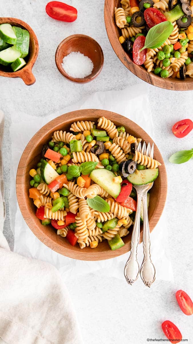 rainbow pasta salad and silverwares on a wood bowl with another bowl of salad and salt, cucumber, and cherry tomatoes placed on a white cloth.