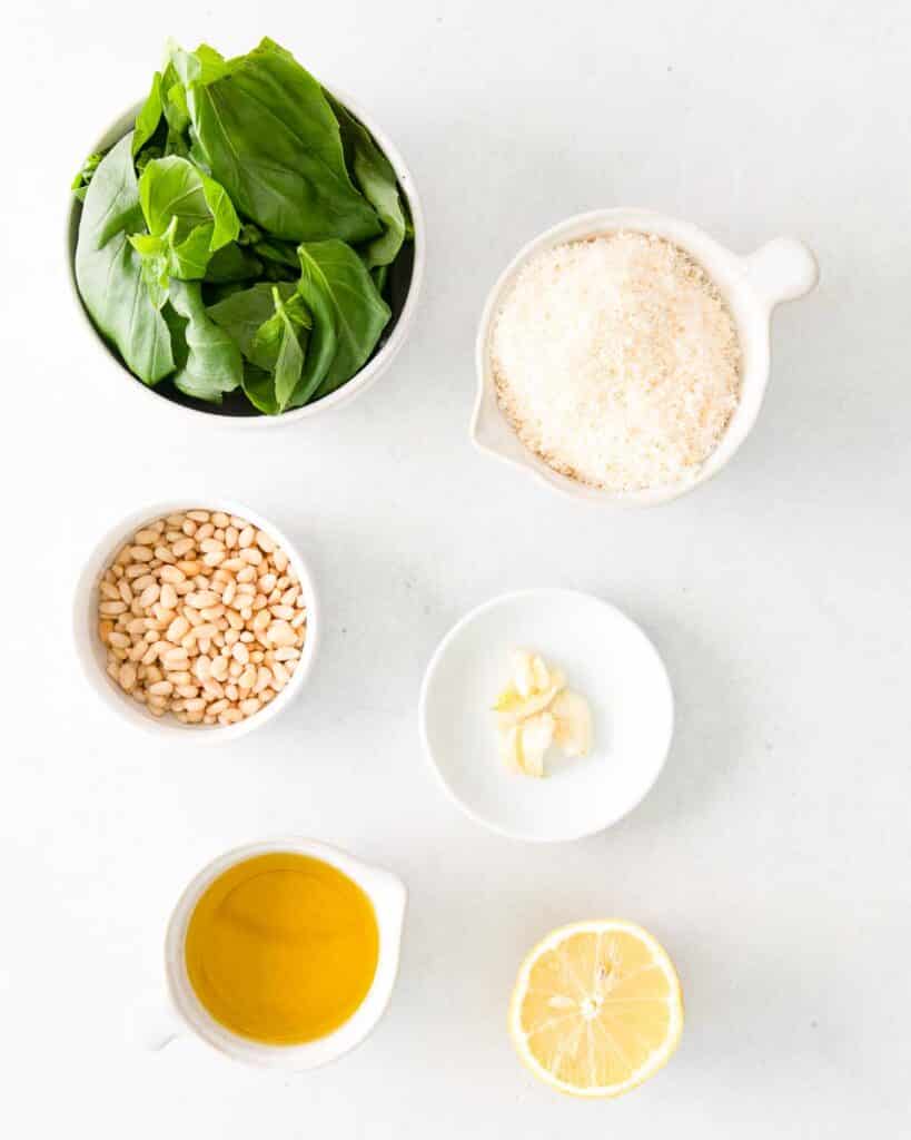 ingredients for pesto sauce which contains basil leaves, parmesan cheese, pine nuts, garlic, olive oil, and a slice of lemon.