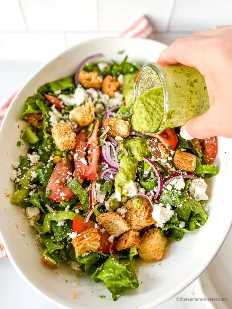 a hand holding a cup of pesto vinaigrette and pouring it in a bowl of salad.