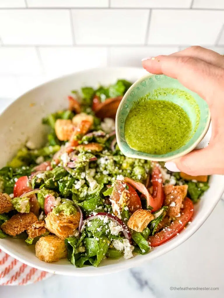 a hand pouring the pesto vinaigrette in a bowl of salad.