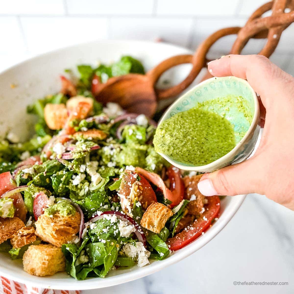a hand holding a small bowl of pesto vinaigrette pouring into a bowl of salad.