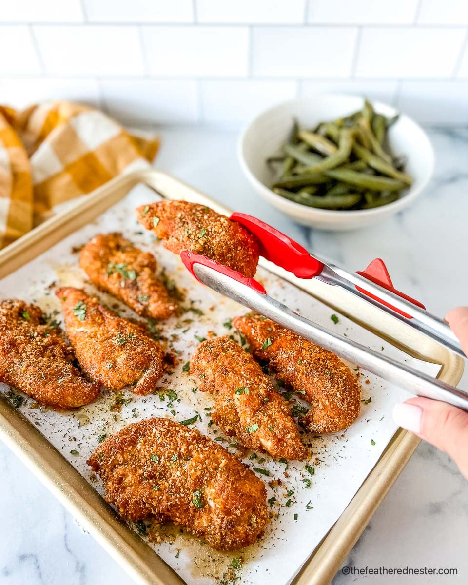 Cooked shake and bake chicken tenders on top of a parchment paper placed in a casserole with a tong holding a piece of crispy chicken tenderloin with a bowl of vegetable and yellow cloth in the background.