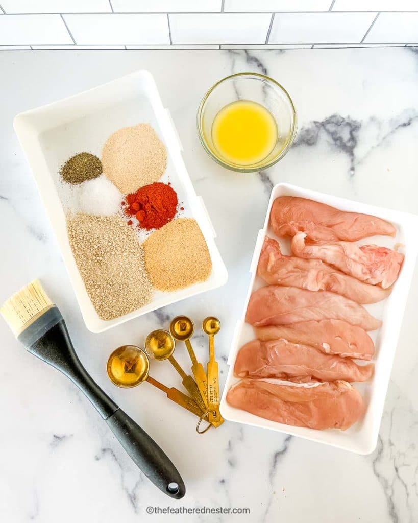 ingredients for shake and bake chicken recipe - melted butter, plate of dried spices, and platter of chicken tenderloins with basting brush and measuring spoon on the side.