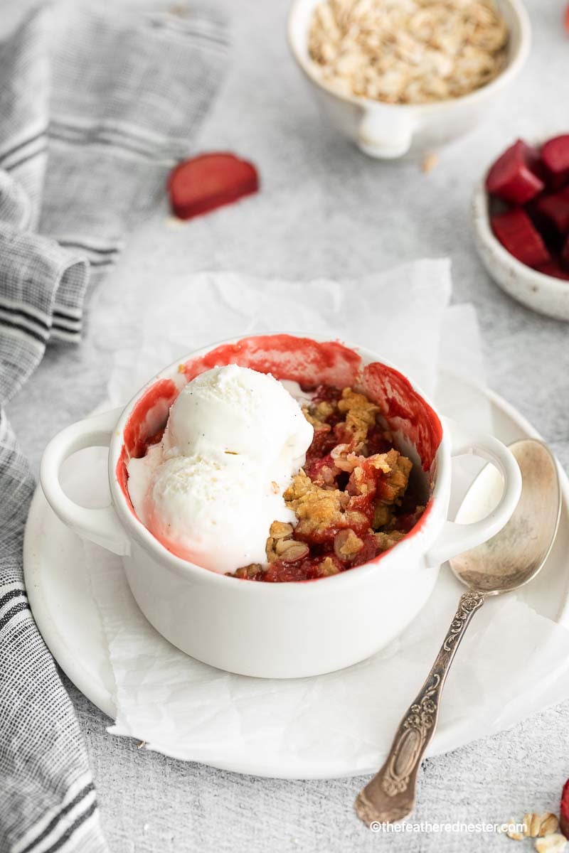 a ramekin with apple strawberry crisp with vanilla ice cream on top and a spoon beside it on top of a plate with parchment paper with gray cloth, bowl of oats and rhubarb in the background.