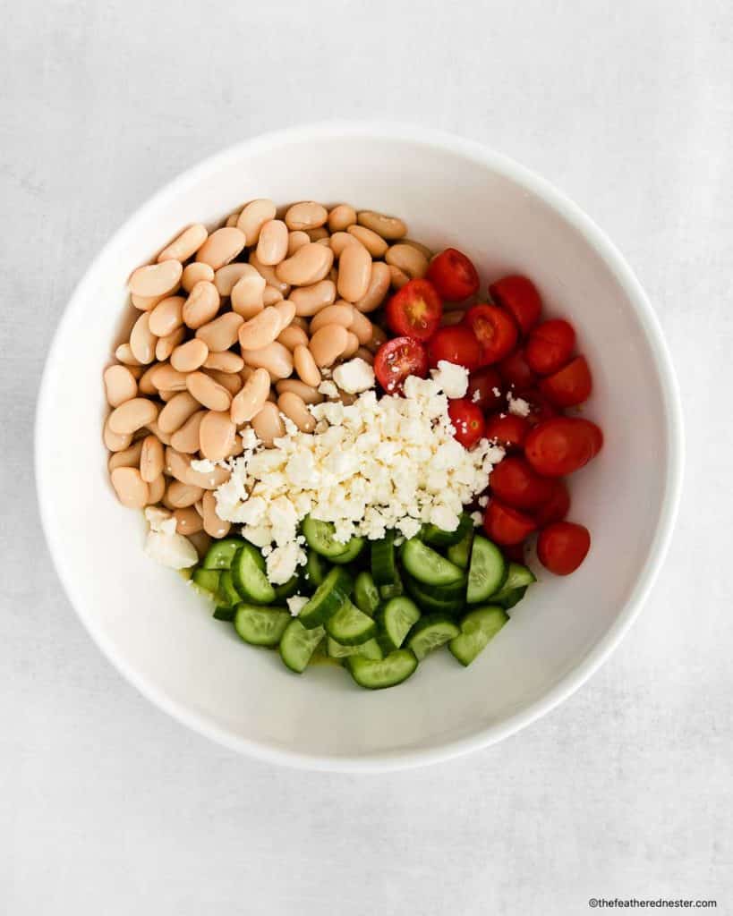 cannellini beans, cherry tomatoes, cucumbers, and feta cheese all placed in a white bowl