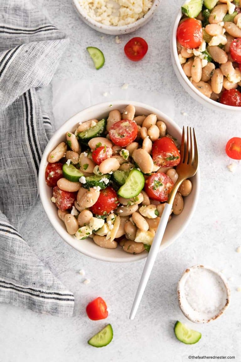 Tuscan white bean salad with a fork in a white bowl with cloth, another bowl of tuscan salad, and chopped tomatoes ad cucumbers at the background