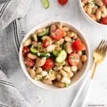 a white bowl of tuscan bean salad with silverware to the right and a striped napkin on the left with the corner of another bowl of white bean salad showing on the top right