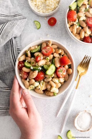 a white bowl of cannellini bean salad with silverware to the right and a striped napkin on the left with the corner of another bowl of white bean salad showing on the top right
