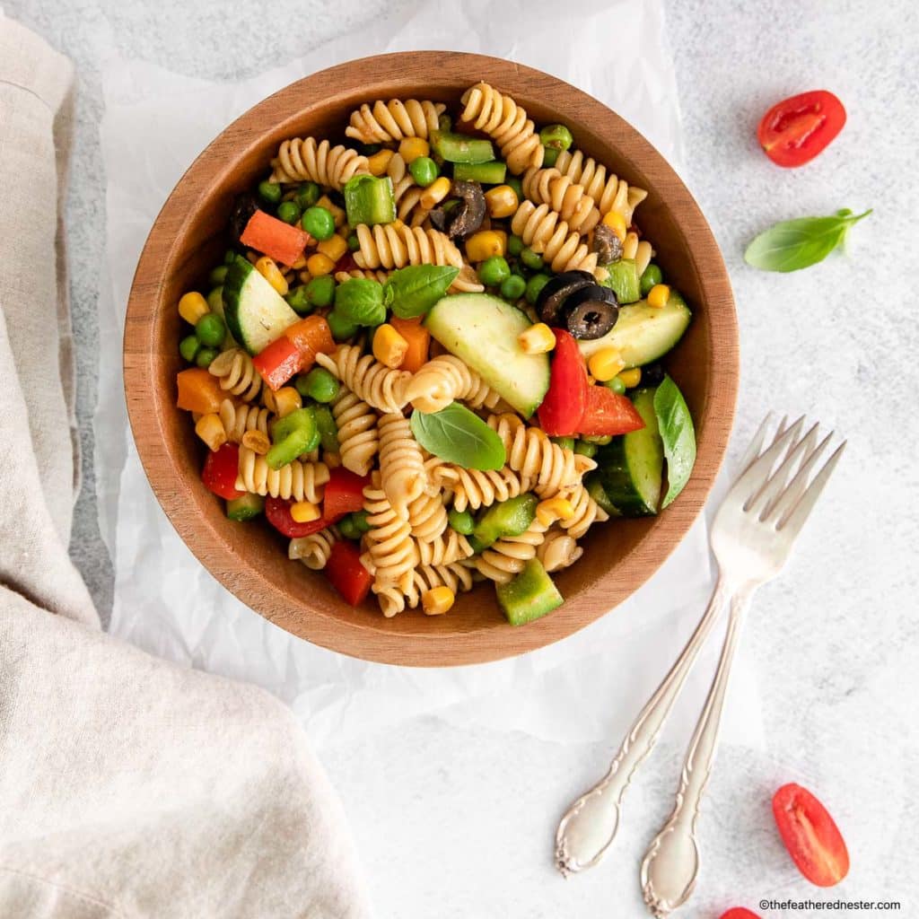 a wooden bowl of pasta salad and salt, cucumber, and cherry tomatoes placed on a white cloth