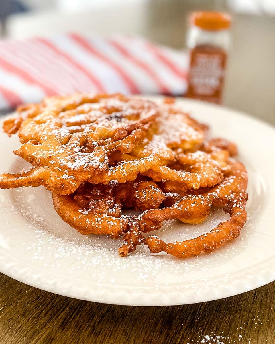 a white plate with Bisquick funnel cake and a red striped cloth and a cinnamon powder bottle at the back.